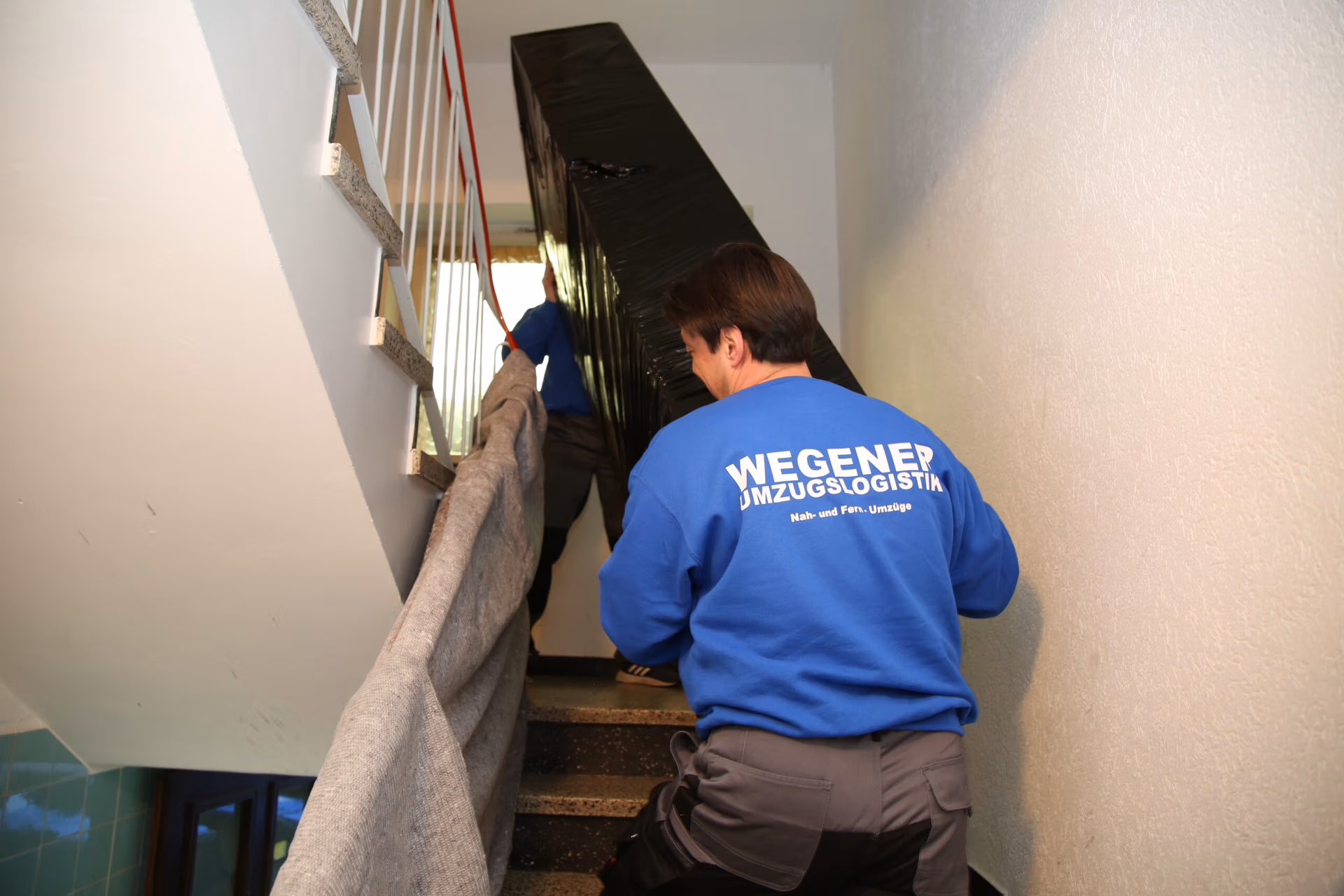 Mitarbeiter der Umzugsfirma Wegener Umzugslogistik tragen einen professionell für den Transport verpackten Schrank in einem engen Treppenhaus.